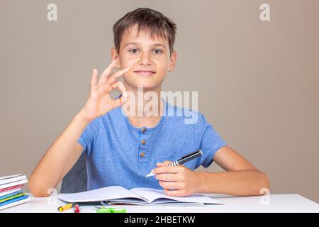 Boy hand holding pen in left hand and writing in a notebook, doing homework. Smiling happy kid enjoy learning and show OK hand gesture. Left Handers Stock Photo