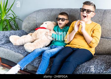 Cute boys, bothers watching movie with 3d glasses, sitting on the sofa in living room, together with teddy bear toy