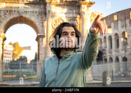 Beautiful man poses for a photo in front of the Arch of Titus. The man with long hair is surprised to see something in front of him and points to it. Stock Photo