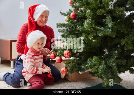 cheerful grandmother and her cute granddaughter girl decorate the Christmas tree. A grandmother and a small child are having fun near a tree indoors. Stock Photo