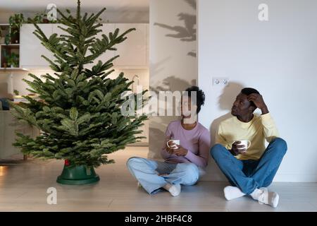 African couple sitting on floor near real Christmas tree, have rest while decorating home for xmas Stock Photo