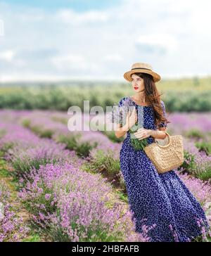 Brunette long-haired woman in a lavender field, in a long blue dress and hat outdoors on a summer day. Stock Photo