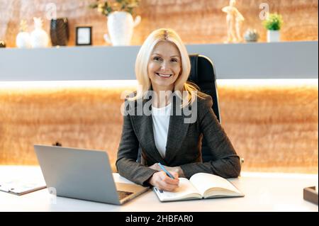 Portrait of beautiful elegant successful middle aged caucasian woman, business lady, corporate top manager, in suit, sitting at the table in the office, looking at the camera, smiling friendly Stock Photo