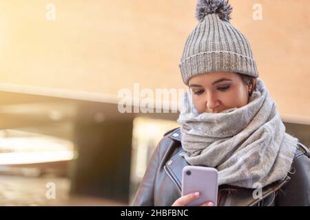 Young millennial wearing knit cap, scarf and leather jacket, while reading a message on cell phone, standing on city street. Stock Photo