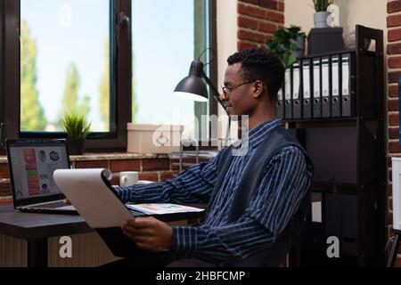 Smiling small business owner looking at laptop screen with analytics data holding clipboard sitting at desk. Entrepreneur with glasses comparing business charts happy with results. Stock Photo