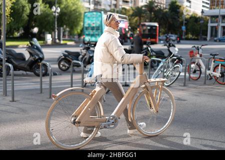 Full body woman in trendy outerwear pushing lumber bike and walking near parking lot on sunny day on street of modern city. Stock Photo