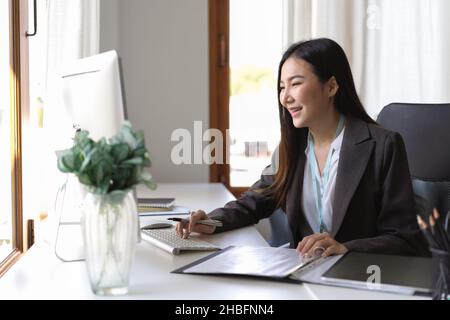 Young asian finance market analyst working at the office on desktop computer. Business woman analyse document on her desk. Stock Photo
