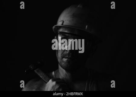 Miner, dirty in coal soot, wearing helmet, protective glasses and with hammer in his hands in black and white photo Stock Photo