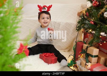 Happy little boy in reindeer antlers headband opening Christmas presents he found under tree Stock Photo