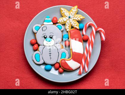 gingerbread cookies decorated with glaze in shape of bear in hat, lollipop, snowflakes on plate on red background Top view Flat lay Christmas treats, Stock Photo