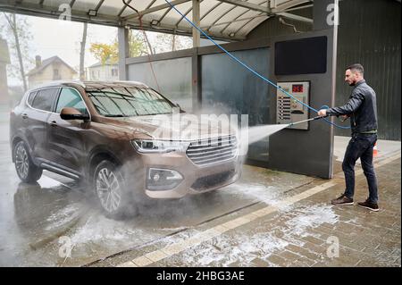 Young man washing car on carwash station outdoor. Handsome worker cleaning automobile, using high pressure water. Stock Photo