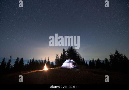 Tourist man sitting in his illuminated tent set up on mountain hill under starry sky on summer night next to campfire. Trees, mountain outlines, sky light on the background. Concept of night camping. Stock Photo