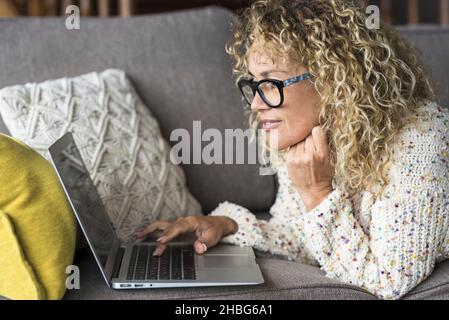 Adult young woman surf the web and use laptop computer laying comfortable on the couch at home. Modern connected people and wireless connection. Conce Stock Photo