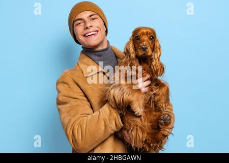 Young handsome happy man, student wearing warm winter clothes and hat standing with cute dog on blue studio background. Stock Photo