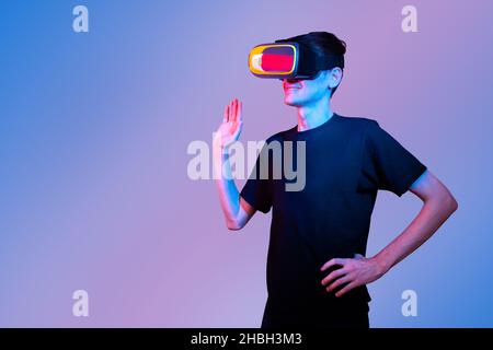 Young man in VR glasses met a friend in virtual reality and waves his hand in greeting. Communication in virtual reality. Neon colors, copy space Stock Photo