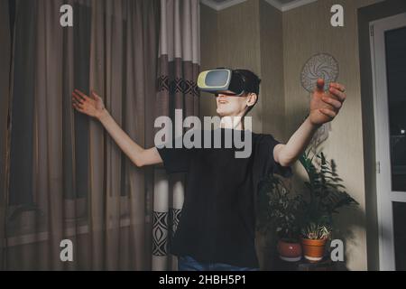 Young man wearing VR glasses met a friend in virtual reality and open his hands in greeting in the home interior. Communication in virtual reality. Stock Photo