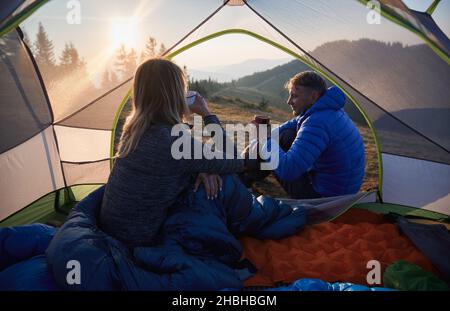 Back view of young woman hiker sitting inside tourist tent and drinking coffee while man resting outside the tent. Couple travelers enjoying morning hot beverage while camping together in mountains. Stock Photo