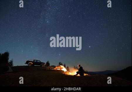 Female camper resting by bonfire and enjoying pleasant atmosphere in camp outdoor. Two tents, wooden table between them and black jeep parked on hill. Beautiful shining stars in the night sky. Stock Photo