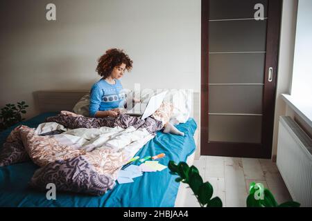 Caucasian curly woman doing online lessons from the bed using laptop in a blue pajama. Quarantine lifestyle. Stock Photo