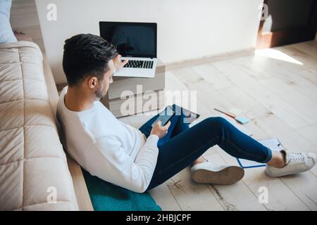 Caucasian man using his laptop while sitting on the floor and texting messages. Remote work. Pandemic Stock Photo