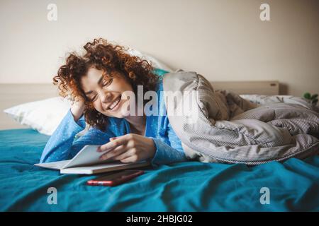 Caucasian woman reading something during leisure time in bedroom. Smiling face. Coronavirus Stock Photo
