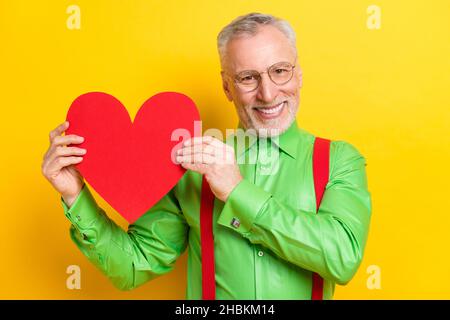 Photo portrait old man smiling in spectacles showing red heart shaped postcard on date isolated vivid yellow color background Stock Photo