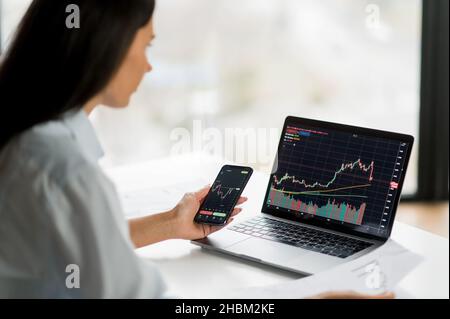 Young adult confident female investor, broker or crypto trader, using laptop and smartphone, buys and sells crypto coins, analyzes charts of trading in stock market and digital cryptocurrency exchange Stock Photo