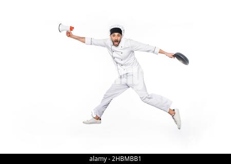 surprised young chef in hat and uniform levitating while holding frying pan and megaphone isolated on white Stock Photo