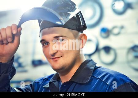 Portrait of Metal Locksmith. Young Caucasian man looks into camera from under visor of protective mask. Real worker in workshop. Stock Photo