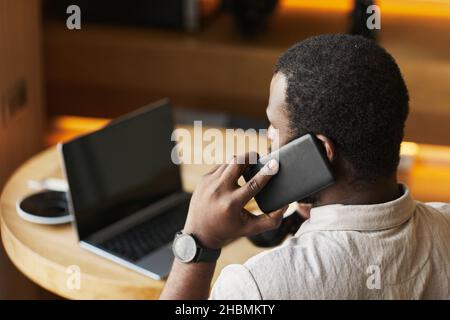 Back view portrait of adult black man speaking by smartphone while working at cafe table in wooden interior, copy space Stock Photo