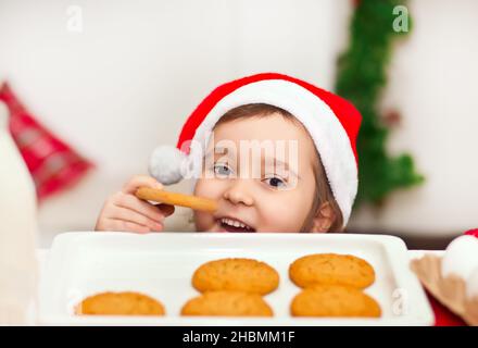 Kid little cute girl in Santa cap taking try to steal cookie from table Stock Photo