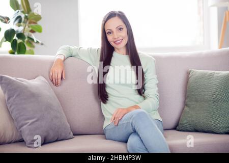 Photo of cute millennial brunette lady sit on sofa wear blue shirt jeans at home alone Stock Photo