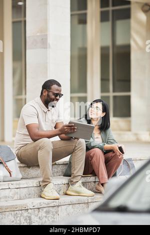 Vertical full length portrait of two young business people chatting while sitting on steps in city Stock Photo