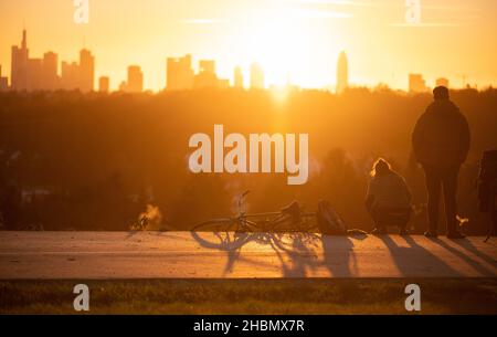 20 December 2021, Hessen, Frankfurt/Main: Two men stand and sit in front of the sunset behind the skyline. Photo: Sebastian Gollnow/dpa Stock Photo