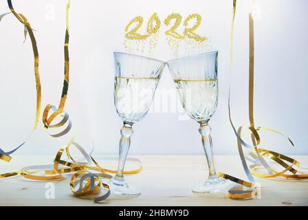 Two champagne glasses toast to new year between golden streamers, bubbles shape the number 2022, light background, selected focus, narrow depth of fie