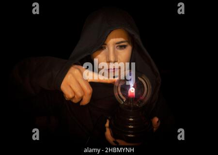 Mysterious Caucasian woman in the 40s with alternative look wearing a hood and piercing on her nose holding plasma lamp touches with index finger appr Stock Photo