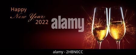 Happy New Year 2022 text, party time with two champagne glasses and fireworks of sparklers against a dark red background with copy space in panoramic Stock Photo
