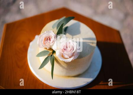 White wedding cake with roses and green leaves, stands on a wooden table. Top view Stock Photo