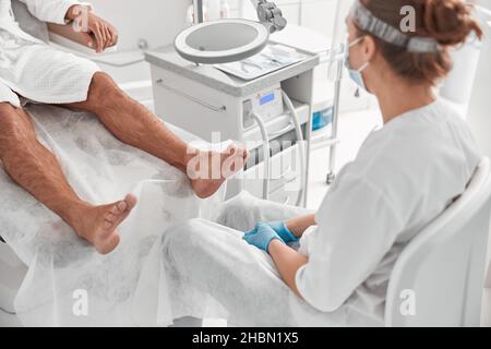 Master of pedicure in white uniform and mask sits near barefoot client in beauty salon Stock Photo