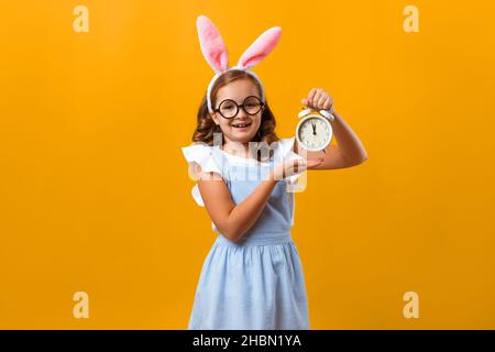 Happy little girl in Easter bunny ears on isolated yellow background. The child is holding an alarm clock. Stock Photo