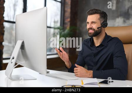 Cheerful guy using handsfree headset and computer to talking online at his workplace. Confident man sitting at the office desk and working with pleasant smile Stock Photo