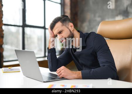 Doubtful caucasian guy sitting at the table at the office. Frustrated pensive man worried about work issues, covering forehead with hand, thinking about troubles and problems Stock Photo