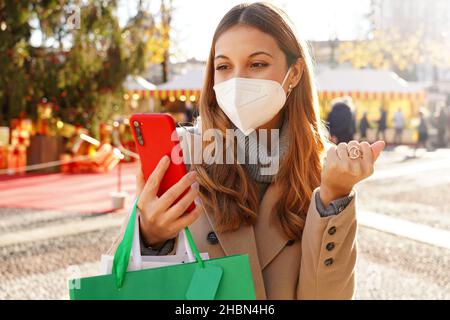 Excited young woman wearing medical mask FFP2 KN95 watching smartphone and holding shopping bags walking in the Christmas Markets Stock Photo