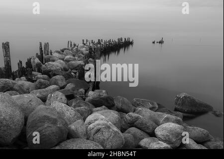 an old pier made of stones and wooden legs is left with metal screeds and the water surface is calm Stock Photo