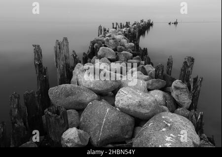 an old pier made of stones and wooden legs is left with metal screeds and the water surface is calm Stock Photo