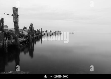 an old pier made of stones and wooden legs is left with metal screeds and the water surface is calm Stock Photo