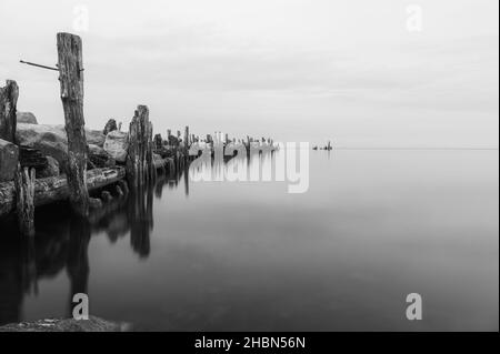 an old pier made of stones and wooden legs is left with metal screeds and the water surface is calm Stock Photo