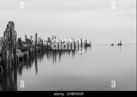 an old pier made of stones and wooden legs is left with metal screeds and the water surface is calm Stock Photo