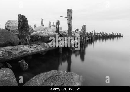 an old pier made of stones and wooden legs is left with metal screeds and the water surface is calm Stock Photo