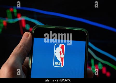 In this photo illustration the American National Basketball Association (NBA) men's professional basketball league logo seen displayed on a smartphone with an economic stock exchange index graph in the background. Stock Photo
